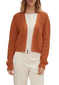 Rustfarbenes Strickjacke mit strukturierten Oberflächen, offener Front und gesenkten Schultern. Getragen über einem hellbeigen Oberteil und kremfarbigen Hosen.