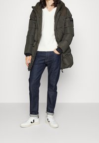 Oliwkowa kurtka puchowa z kapturem, zapinana na zamek, z naszywkami, noszona na białym swetrze V-neck i ciemnoniebieskich dżinsach, dopełniona białymi trampkami.