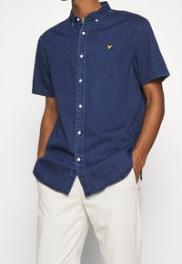 Camicia in denim a maniche corte di colore blu scuro, con chiusura a bottoni sul davanti, colletto casual, bottoni bianchi e ricamo del logo giallo sul petto.