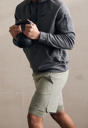 Person in grauer Kapuzenjacke und beigen Shorts hält eine schwarze Kettlebell mit beiden Händen in einer Fitnesspose vor einem schlichten Hintergrund.