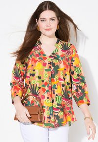 Blusa a fantasia di frutta colorata con scollatura a V, maniche lunghe e bottoni, abbinata a una borsa marrone. I pantaloni bianchi completano l'outfit.