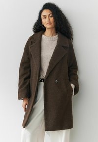 Brun oversized kappa i en texturerad tyg, med dubbelknäppt design, stora slag och framfickor, bärs över en lätt tröja.