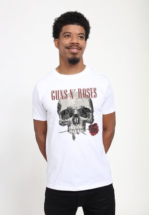Mężczyzna uśmiecha się, nosi białą koszulkę Guns N' Roses z czaszką trzymającą różę w ustach, stoi na jednolitym jasnym tle.
