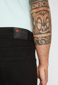 Melna denima bikses ar matētu apdari, ar ādas birku, uz kuras ir uzraksts "DENIM PROJECT", un redzamu aizmugurējo kabatu. Parādīta āda ar detalizētu tetovējumu.