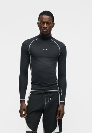 Hombre negro con un rash guard negro ceñido de manga larga y pantalones cortos de tabla negros con ribete blanco, de pie contra un fondo liso.