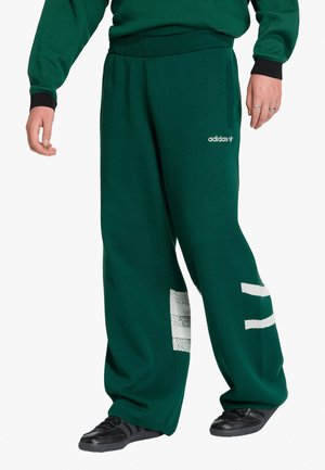 Persona che indossa pantaloni da tuta Adidas verde scuro con strisce bianche e scarpe da ginnastica nere, in piedi con le mani rilassate lungo i fianchi.
