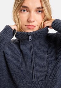 Femme blonde aux yeux clairs portant un pull en maille côtelée bleu marine, tirant sur le col montant zippé avec les deux mains.