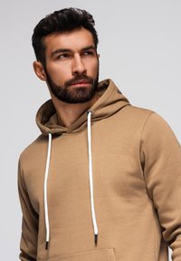 Muž s tmavými vlasy a beardem, který má na sobě béžovou mikinu s bílými tkaničkami, lehce se dívající na stranu proti jednolitému světlému pozadí.