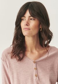Top in morbido tessuto rosa chiaro con scollatura a V e tre bottoni in legno sul davanti. Presenta una vestibilità rilassata e dettagli leggermente usurati.
