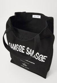 Black canvas tote bag with dual handles, featuring bold white text and a label inside. Simple design with a spacious opening.