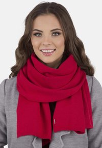 Fular tricotat roșu înfășurat în jurul gâtului, având o textură netedă și o formă largă, rectangulară, asortat cu un palton gri.
