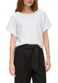 Top en lin blanc à coupe ample et manches larges, associé à un pantalon noir taille haute avec une ceinture nouée.