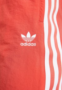 Atletické šortky Adidas v korálové barvě s bílými trojpruhy a vyšívaným logem. Hladká textura s pohodlným střihem.