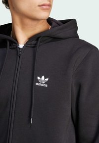 Černá mikina na zip vyrobená z hladkého materiálu, která má bílé logo Adidas na levé části hrudi a stahovací kapuci.