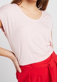Světle růžový top s krátkým rukávem a kulatým výstřihem, spojený s červenými kalhotami s vysokým pasem a detailem zavázání vpředu. Měkká textura, uvolněný střih.