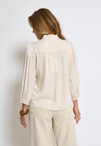 Hellbeige Bluse mit 3/4-Ärmeln, Rüschenausschnitt und gerafften Details. Glatter Stoff mit lockerer Passform, kombiniert mit cremefarbenen Hosen.