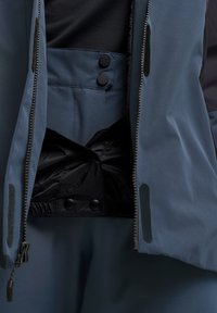 Blaue wasserdichte Jacke mit einem Reißverschluss vorne, schwarzen Akzenten und Druckknöpfen. Das Innere besitzt ein strukturiertes schwarzes Futter und einen elastischen Bund.