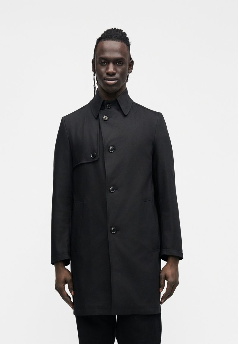 Svart, midjelang kåpe med knappelukking foran, spiss krage og to sidelommer. Jevn tekstur med minimalistiske designelementer.