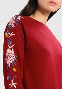 Suéter rojo con patrón floral bordado en la manga izquierda, con detalles en rosa, azul y amarillo. Textura suave, diseño de cuello redondo.