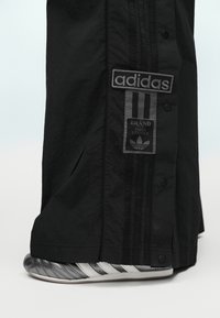 Μαύρο παντελόνι Adidas με κουμπιά τύπου τσίμπημα που καλύπτουν εν μέρει ένα ασημένιο και λευκό αθλητικό παπούτσι Adidas με τρεις ρίγες.