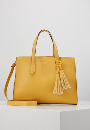 Sac à main en cuir jaune avec double poignées, bandoulière amovible et deux décorations à pompons sur une surface blanche.