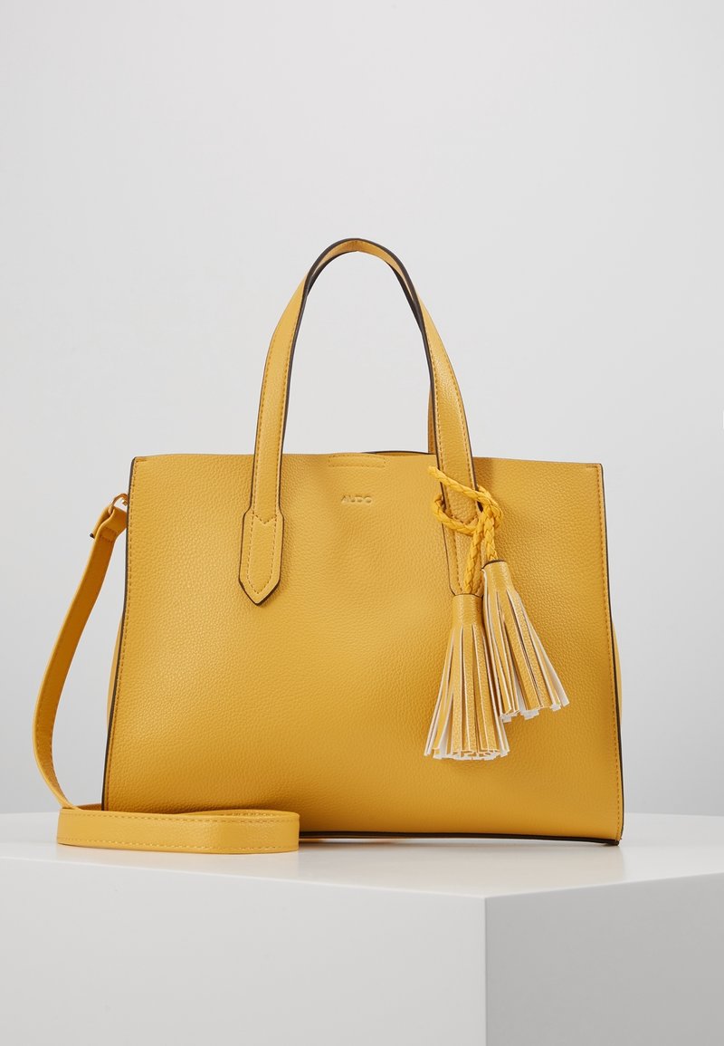 Sac à main en cuir jaune avec double poignées, bandoulière amovible et deux décorations à pompons sur une surface blanche.