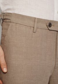 Beige broek met een patroon van geruite structuren, voorzien van een knoop en riemlussen. De stof is lichtgewicht en op maat gemaakt.