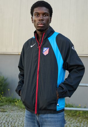 Black lightweight jacket featuring blue sides, red zipper, and football club emblem. Long sleeves with elastic cuffs. Worn over blue jeans.