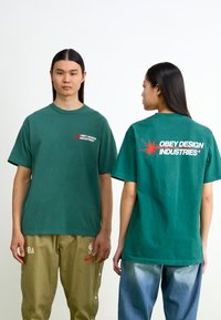 Tricou verde din bumbac cu o croială supradimensionată, logo pe partea stângă a pieptului și un grafic îndrăzneț pe spate. Mâneci scurte și guler rotund.