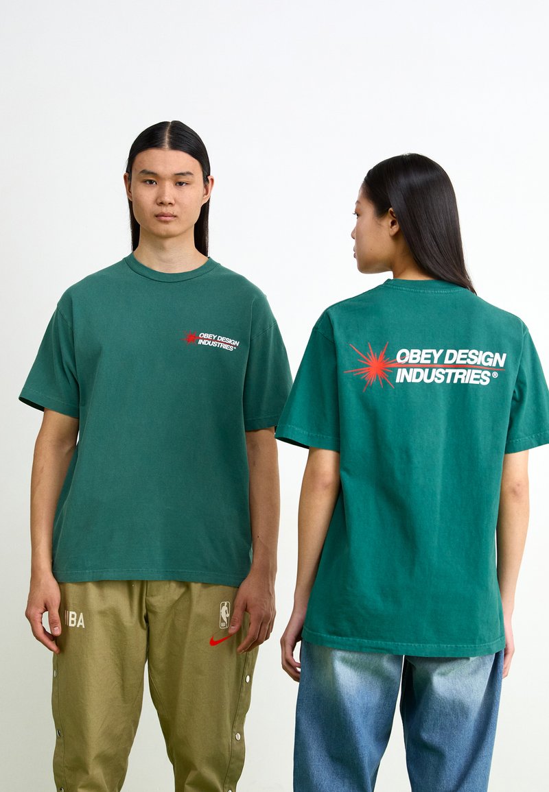 Tricou verde din bumbac cu o croială supradimensionată, logo pe partea stângă a pieptului și un grafic îndrăzneț pe spate. Mâneci scurte și guler rotund.