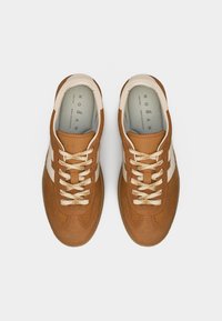 Par de zapatillas de ante marrón con cordones crema y rayas blancas en los laterales, vistas desde arriba sobre un fondo gris claro.