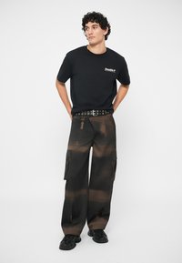 Tricou negru din bumbac cu logo pe piept, asortat cu pantaloni cargo închiși la culoare, cu model tie-dye, buzunare laterale și curea cu ținte.