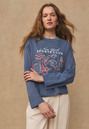 Kobieta z długimi włosami ubrana w niebieską bluzkę z długim rękawem z napisem „New York Sports Club” i kreskówkowymi królikami grającymi w tenisa, w połączeniu z jasnymi spodniami.
