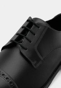 Čierna kožená šnurovacia spoločenská obuv so zatvoreným šnurovaním, hladkým povrchom a dekoratívnymi dierkami blízko špičky na bielom pozadí.