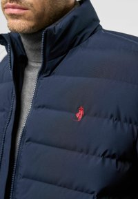 Hombre vistiendo una chaqueta acolchada azul marino con un logotipo rojo bordado sobre un suéter de cuello alto gris, primer plano en el área del pecho y del cuello.