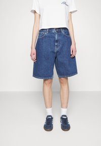 Pantalones cortos de mezclilla azul con un ajuste holgado, longitud hasta la rodilla y dobladillo cosido. Combinados con una camiseta blanca corta y zapatillas deportivas azules.