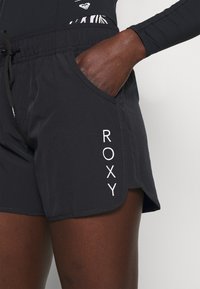 Black swim shorts made of lightweight material, featuring an elastic waistband, side pockets, and white "ROXY" logo.