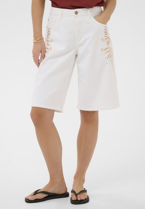 Fehér, térdig érő rövidnadrág oldalsó hímzéssel, bordó felsővel és fekete papucsokkal viselve, laza, hétköznapi stílust mutatva.