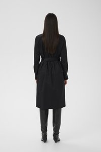 Hosszú egyenes hajú nő, fekete, övvel ellátott, hosszú ujjú ruhában és térdig érő fekete bőr csizmában, háttal állva.