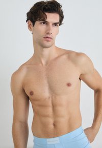 Svetlo modri bokseric s belim pasom, na katerem je logo "KARL LAGERFELD". Model ima kratke valovite lase in tonirano telo.