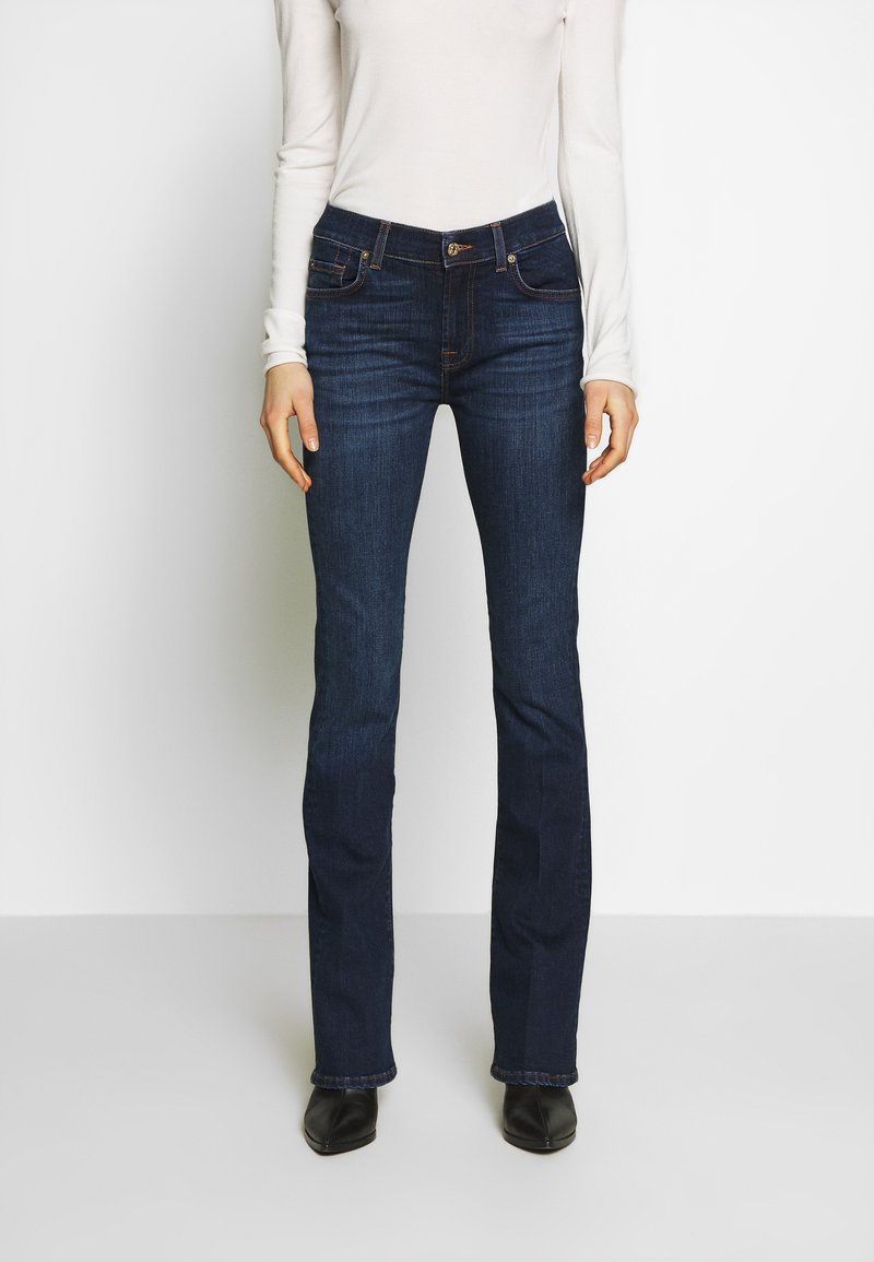 Donkerblauwe flared jeans van denim; met voorzakken en knoopsluiting; gecombineerd met een nauwsluitend wit longsleeve shirt.