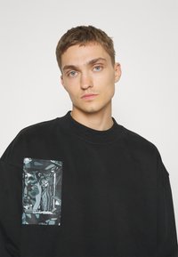 Zwarte sweatshirt met een ronde hals, met een groot grafisch zakontwerp in tinten blauw en grijs aan de linkerkant.