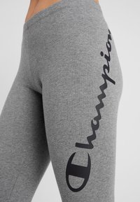 Szare legginsy w stretchu z dopasowanym krojem. Czarny logo biegnie w pionie wzdłuż lewej strony, podkreślając nazwę marki.