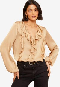 Blouse en satin beige avec un col en V froncé, des manches longues et des poignets froncés, associée à un jean noir taille haute et une ceinture noire.