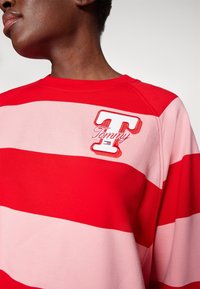 Sudadera a rayas rojas y rosas con cuello redondo. Presenta un logo bordado de la letra "T" y el nombre "Tommy" en el pecho. Textura suave.