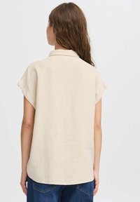 Camicia beige a maniche corte con colletto e vestibilità rilassata, realizzata in tessuto testurizzato, orlata con fondo arrotondato.