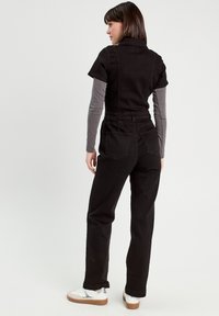 Jumpsuit in denim nero con maniche corte, colletto e tasche posteriori, abbinato a un top grigio a maniche lunghe e sneaker bianche con suole marroni.