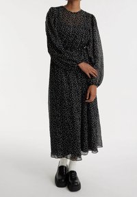 Schwarzes Polka-Dot-Kleid aus transparentem Material, mit langen Ärmeln und tailliertem Bund. Kombiniert mit schwarzen Plateauschuhen und weißen Socken.