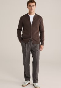 Barna cipzáras kardigánt, fehér pólót, kockás nadrágot és fehér tornacipőt viselő férfi, sima bézs háttér előtt áll.