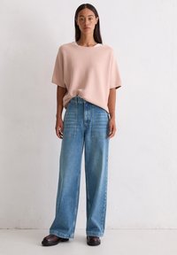 Jasnoróżowy, oversize'owy sweter z szerokim dekoltem, w zestawieniu z luźnymi niebieskimi jeansami z kieszeniami naszytymi oraz prostą nogawką.